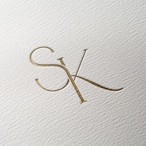 Op de afbeelding: Een gouden monogram met de letters "SK" in een gestileerd handschrift.