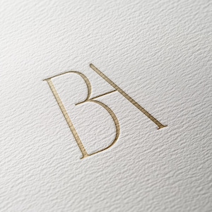 May include: A gold-coloured monogram featuring the letters "BA" in a simple, elegant font.