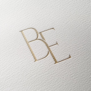 May include: A gold-colored monogram with the letters "BE" in a simple, elegant font.