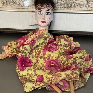 May include: A wooden puppet with a painted face and black hair, wearing a green and pink floral dress. The puppet is sitting on a gray surface with two wooden sticks.