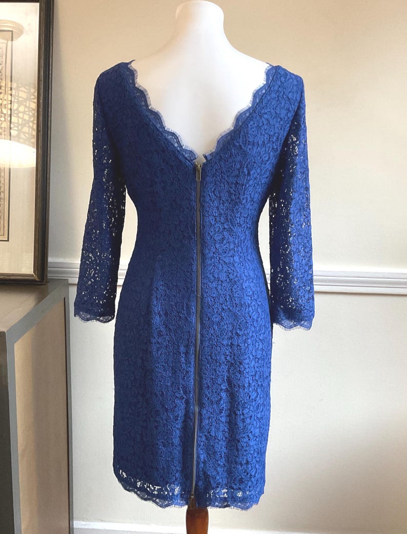 May include: A cobalt blue lace dress with long sleeves and a scalloped neckline. The dress features a full-length zipper down the back and is displayed on a mannequin. The dress is a knee-length style.