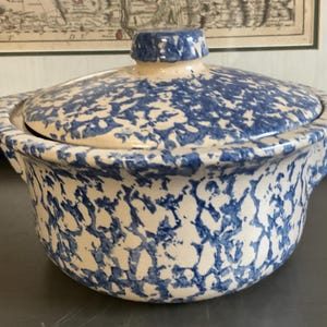 May include: A ceramic casserole dish with a lid. The dish and lid are off-white with a blue speckled pattern. The dish has two handles and a knob on the lid. The dish is likely vintage or antique.