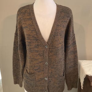 May include: A brown and gray marled knit cardigan sweater with a V-neck, button closure, and two front pockets. The sweater is displayed on a mannequin.