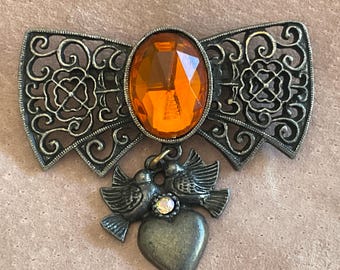 May include: A silver-toned brooch in the shape of a bow with a large orange faceted glass stone in the center. The bow has a delicate filigree design. Hanging from the bow are two silver-toned birds with a heart-shaped charm.