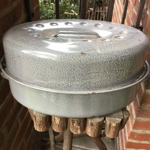 May include: A vintage, speckled gray enamelware roaster with a lid and handle. The roaster sits on a rustic wooden stand with a brick background. The roaster is round and has a handle on the lid.
