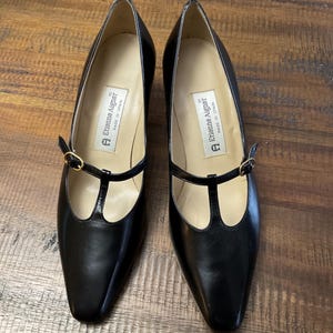 May include: A pair of black T-strap pumps with a rounded toe and a thin gold-tone buckle. The interior features a white label with the text Etienne Aigner and Made in Spain printed in black. The shoes have a smooth finish and a low heel profile.