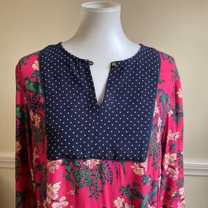 May include: A long-sleeved tunic with a vibrant floral and bird print in shades of pink, green, and yellow. The neckline features a navy blue panel with small yellow polka dots. A small gold-colored button accents the neckline.