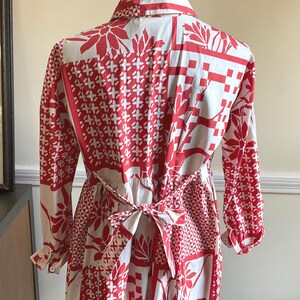 May include: A long-sleeved shirt featuring a white base with a vibrant red patchwork-style print. The pattern includes various geometric shapes, floral motifs, and grid designs. The shirt has a collar and a tie-back waist detail with a bow.