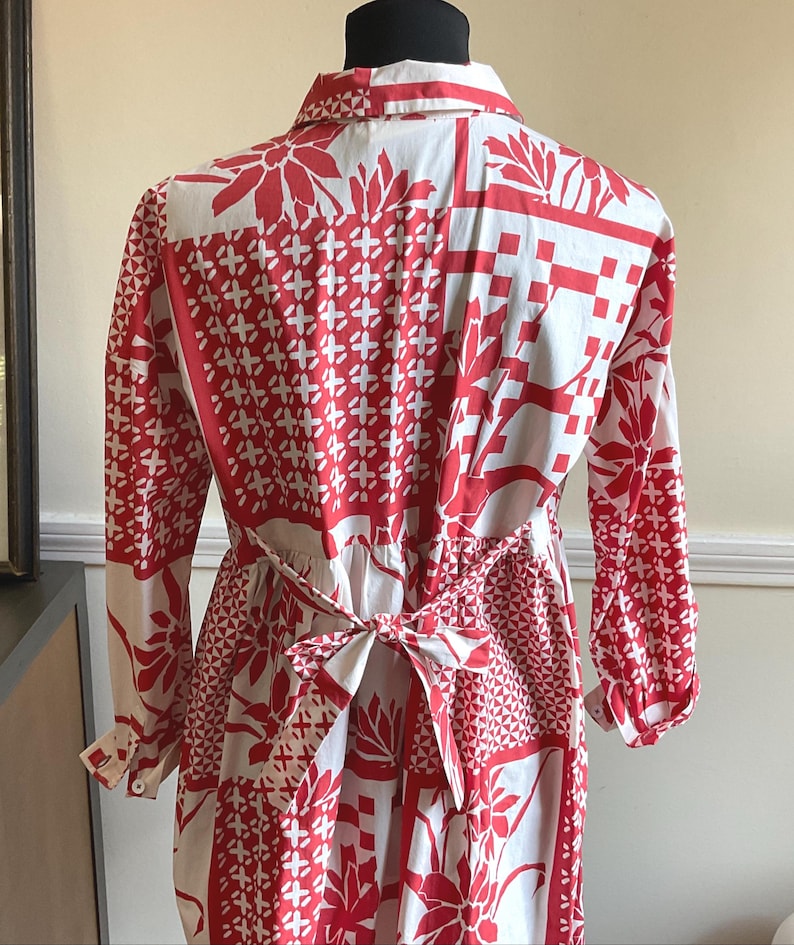 May include: A long-sleeved shirt featuring a white base with a vibrant red patchwork-style print. The pattern includes various geometric shapes, floral motifs, and grid designs. The shirt has a collar and a tie-back waist detail with a bow.