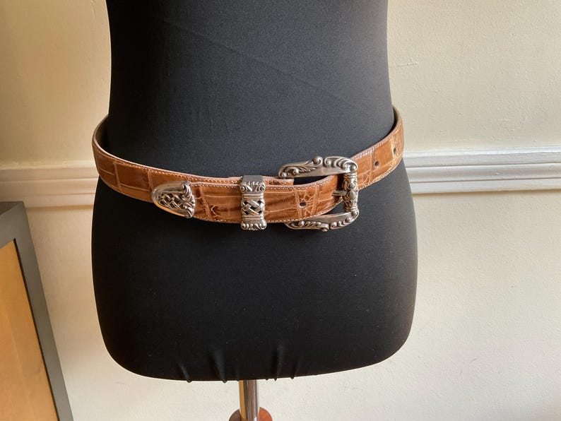 Puede incluir: Un cintur&oacute;n de color tostado con un patr&oacute;n de textura de piel de cocodrilo que presenta una hebilla de metal plateado ornamentada, una trabilla de metal plateado a juego y una punta de metal plateado decorativa, sujeto alrededor de un torso de maniqu&iacute; negro.