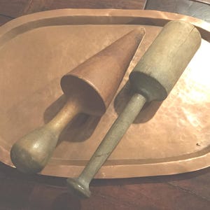 May include: Two antique wooden kitchen tools rest on a copper tray. One tool is cone-shaped, the other is cylindrical. The tray has a rounded rectangular shape. The tools and tray have a rustic, vintage appearance.