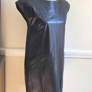 May include: A black leather dress with a round neckline and cap sleeves. The dress has a straight silhouette and a single pocket on the left side. The dress is displayed on a white mannequin.
