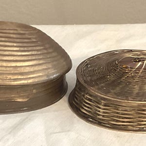 May include: Two decorative boxes. One is shaped like a clam shell, with a ridged, bronze-colored lid. The other is a woven basket design with a matching lid. Both are on a white surface.
