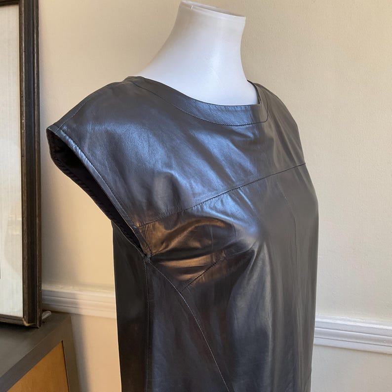 May include: A grey leather dress with cap sleeves. The dress features a square neckline and is displayed on a mannequin. The leather has a smooth texture and a subtle sheen. The dress is a solid colour and has a tailored fit.