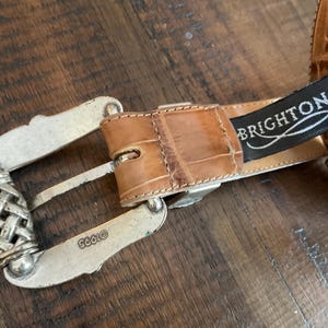 Puede incluir: Un cintur&oacute;n color canela con un patr&oacute;n texturizado en relieve y una hebilla de metal plateado. La hebilla presenta un detalle de metal trenzado y est&aacute; grabada con el texto 1995. Una etiqueta de tela negra con el texto blanco BRIGHTON est&aacute; unida.