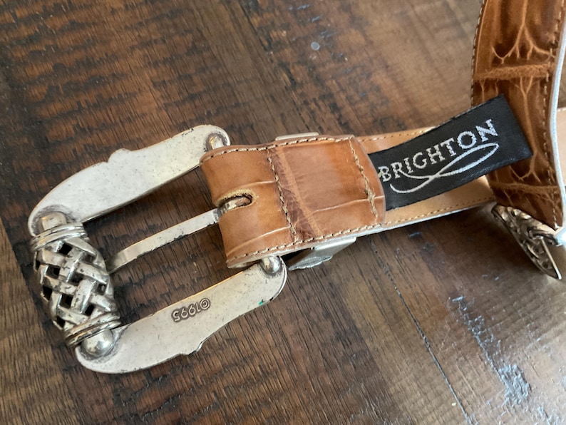 Puede incluir: Un cintur&oacute;n color canela con un patr&oacute;n texturizado en relieve y una hebilla de metal plateado. La hebilla presenta un detalle de metal trenzado y est&aacute; grabada con el texto 1995. Una etiqueta de tela negra con el texto blanco BRIGHTON est&aacute; unida.