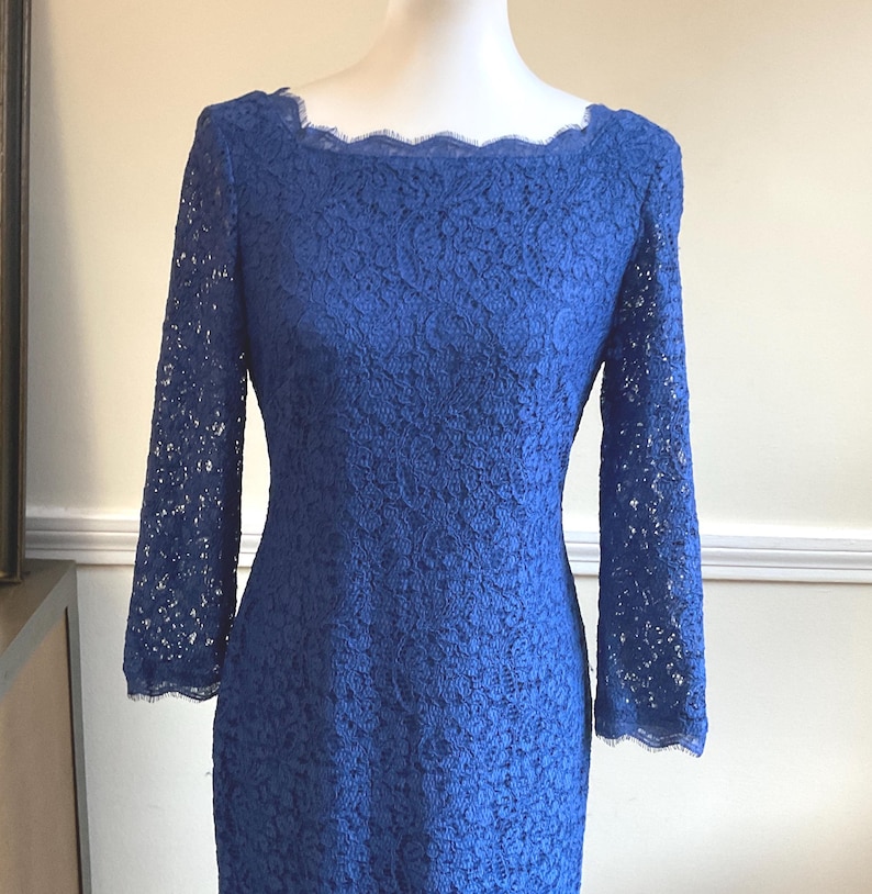 May include: A royal blue lace dress with long sleeves and a scalloped neckline. The dress is displayed on a mannequin, showcasing its intricate floral lace pattern. The sleeves extend to the mid-forearm.