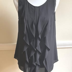 May include: A sleeveless black blouse featuring a round neckline and a vertical row of cascading ruffles down the center front. The garment is made of a lightweight, semi-sheer fabric and includes a single small button closure at the top center.