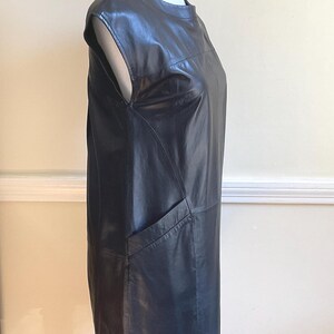 May include: A black leather dress with a round neckline and cap sleeves. The dress has a straight silhouette and a single pocket on the front. The dress is displayed on a white mannequin.