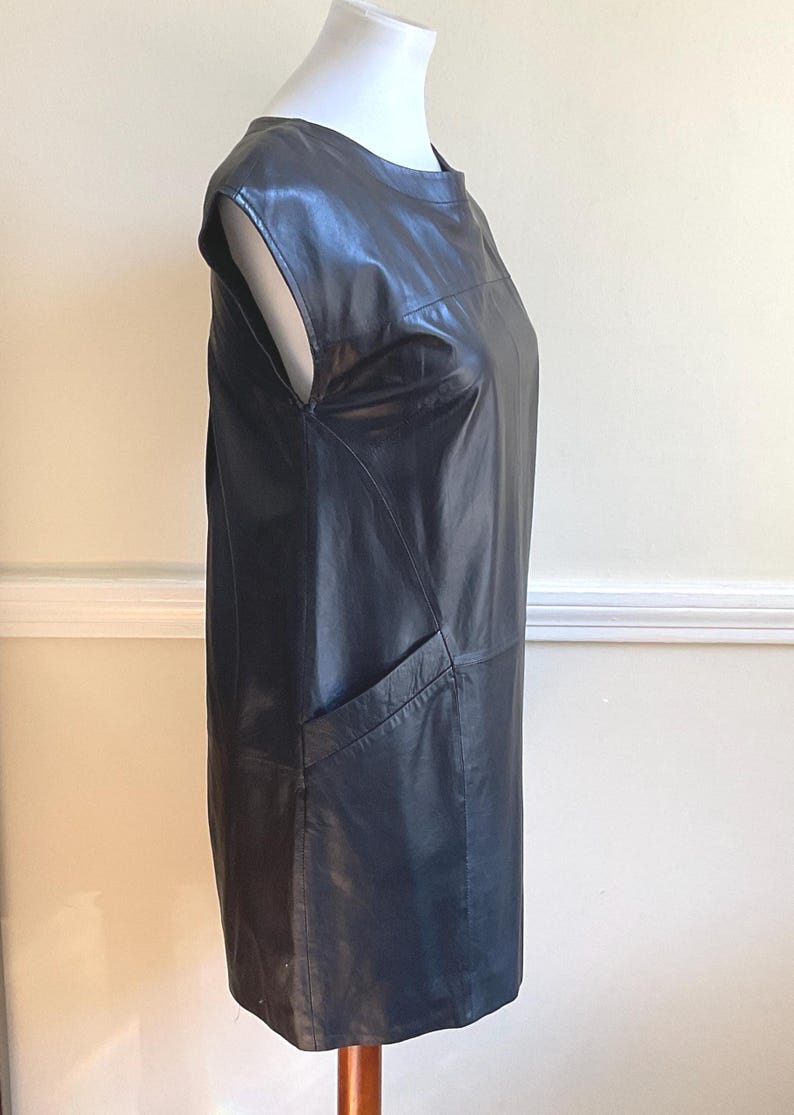 May include: A black leather dress with a round neckline and cap sleeves. The dress has a straight silhouette and a single pocket on the front. The dress is displayed on a white mannequin.