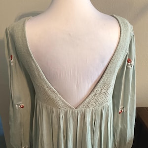K&ouml;nnte beinhalten: Hellgr&uuml;nes Kleid mit tiefem R&uuml;ckenausschnitt und langen &Auml;rmeln. Das Kleid ist mit kleinen roten und wei&szlig;en Blumenstickereien verziert. Der Ausschnitt und der obere R&uuml;cken haben ein strukturiertes Webmuster. Der Stoff hat eine subtile, plissierte Textur.