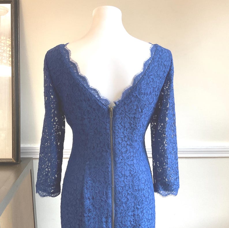 May include: A long-sleeved, navy blue lace dress with a scalloped neckline and a full-length zipper down the back. The dress is displayed on a mannequin, showcasing its intricate floral lace pattern.