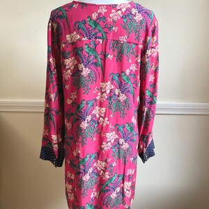 May include: A long-sleeved, pink dress with a vibrant floral and bird pattern. The design features green birds, white flowers, and purple accents. The cuffs are navy blue with white polka dots. The dress is displayed on a mannequin.