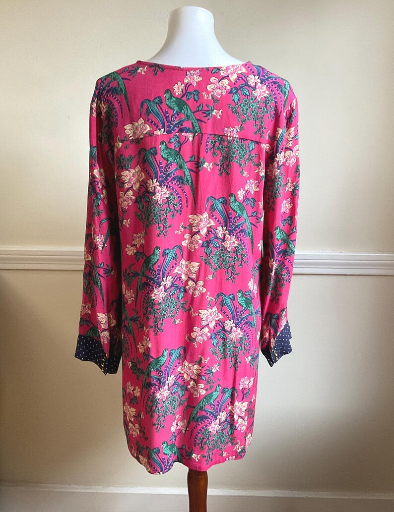 May include: A long-sleeved, pink dress with a vibrant floral and bird pattern. The design features green birds, white flowers, and purple accents. The cuffs are navy blue with white polka dots. The dress is displayed on a mannequin.