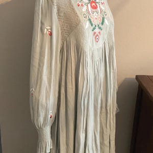 K&ouml;nnte beinhalten: Hellgr&uuml;nes Kleid mit langen &Auml;rmeln und floraler Stickerei. Das Kleid hat einen runden Ausschnitt, Spitzendetails und eine geraffte Taille. Die &Auml;rmel haben eine ger&uuml;schte Manschette. Die Stickerei umfasst rote, wei&szlig;e und gr&uuml;ne Blumenmuster.
