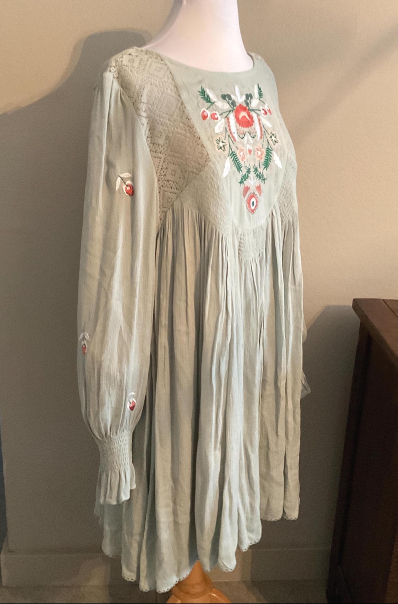 K&ouml;nnte beinhalten: Hellgr&uuml;nes Kleid mit langen &Auml;rmeln und floraler Stickerei. Das Kleid hat einen runden Ausschnitt, Spitzendetails und eine geraffte Taille. Die &Auml;rmel haben eine ger&uuml;schte Manschette. Die Stickerei umfasst rote, wei&szlig;e und gr&uuml;ne Blumenmuster.