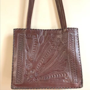 May include: A brown leather tote bag with a floral and leaf design. The bag has two long straps and a rectangular shape. The leather is embossed with a detailed pattern, and the edges are stitched.