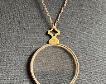 May include: A gold-toned pendant necklace featuring a circular magnifying glass lens encased in a thin metal frame. The pendant is attached to a delicate gold-toned chain by a decorative, cross-shaped bail that bears the stamped letter T.