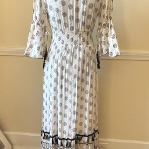 May include: White maxi dress with a black and white floral pattern. The dress features long, flared sleeves, a cinched waist, and a tiered skirt with black tassels. The dress is displayed on a mannequin.