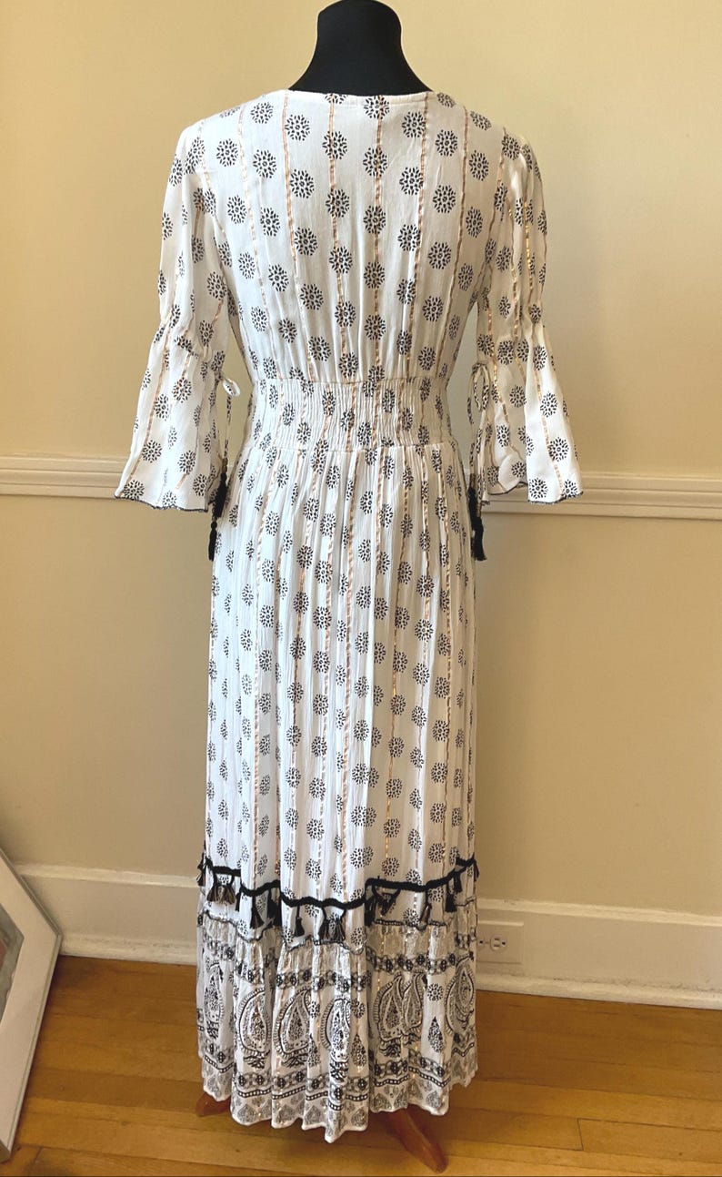 May include: White maxi dress with a black and white floral pattern. The dress features long, flared sleeves, a cinched waist, and a tiered skirt with black tassels. The dress is displayed on a mannequin.