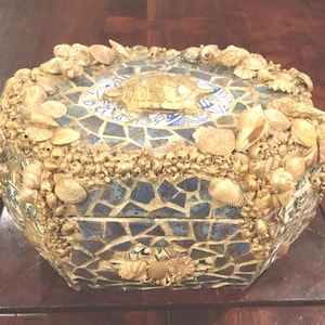 May include: A decorative box with a mosaic design featuring blue and white tiles. The box is adorned with seashells and a gold-colored turtle on the lid. The box is a unique piece of home decor.