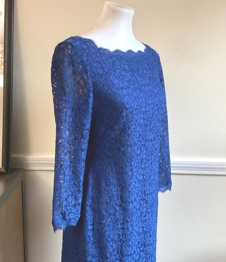 May include: A royal blue lace dress with long sleeves and a scalloped neckline. The dress is displayed on a mannequin, showcasing its intricate floral lace pattern. The dress is a classic style, suitable for formal occasions.