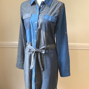 May include: A denim shirt dress in shades of blue and gray, featuring a button-down front, two chest pockets, and a matching tie belt. The dress has long sleeves and is displayed on a mannequin.