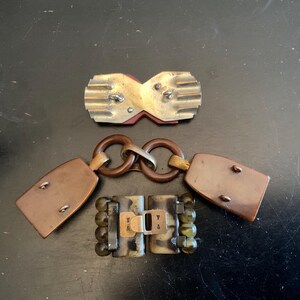 May include: Three vintage metal and plastic belt buckles. The first buckle is a silver-toned metal buckle with a red and white design. The second buckle is a brown plastic buckle with a gold-toned metal ring. The third buckle is a brown plastic buckle with a gold-toned metal clasp and green and brown beads.