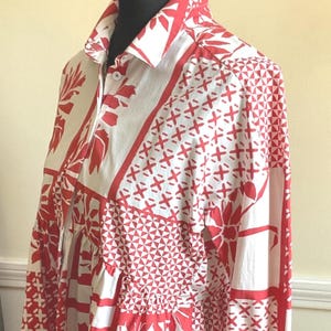 May include: A long-sleeved button-down shirt featuring a white base with a bold red geometric and floral patchwork pattern. The garment has a classic collar, a front button placket with visible white buttons, and a structured fit on a mannequin.