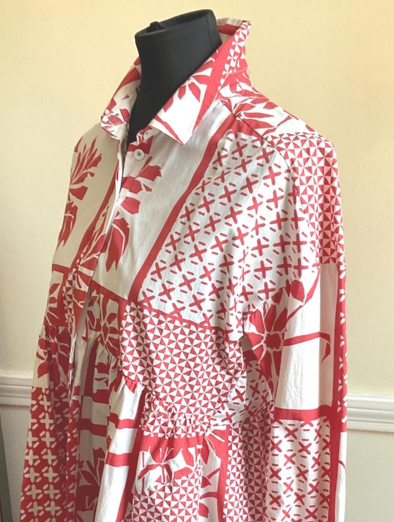 May include: A long-sleeved button-down shirt featuring a white base with a bold red geometric and floral patchwork pattern. The garment has a classic collar, a front button placket with visible white buttons, and a structured fit on a mannequin.
