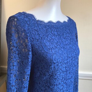 May include: A royal blue lace top with long sleeves and a scalloped neckline. The intricate floral lace pattern covers the entire garment. The top is displayed on a mannequin.
