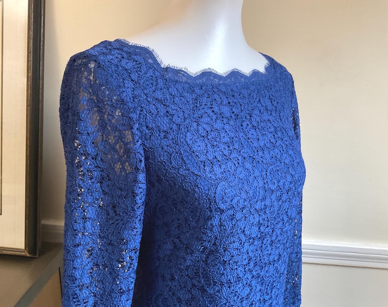 May include: A royal blue lace top with long sleeves and a scalloped neckline. The intricate floral lace pattern covers the entire garment. The top is displayed on a mannequin.