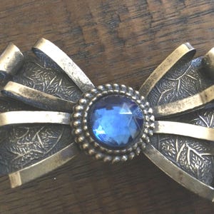 May include: A silver-toned bow brooch with a blue rhinestone in the center. The bow is made of multiple metal strips with a leaf pattern.