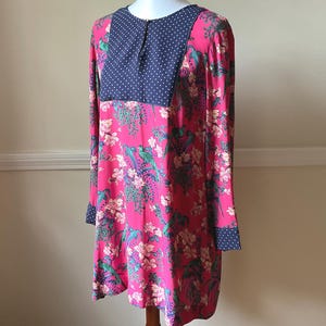 May include: A long-sleeved dress with a vibrant pink floral pattern, featuring a navy blue polka dot yoke and cuffs. The dress is displayed on a mannequin, showcasing its design and fit. The dress has a round neckline.