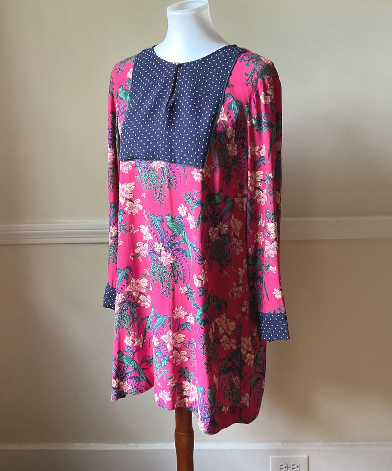 May include: A long-sleeved dress with a vibrant pink floral pattern, featuring a navy blue polka dot yoke and cuffs. The dress is displayed on a mannequin, showcasing its design and fit. The dress has a round neckline.