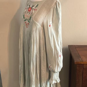 K&ouml;nnte beinhalten: Hellgr&uuml;nes Kleid mit floraler Stickerei und langen &Auml;rmeln. Das Kleid hat einen runden Ausschnitt und einen gerafften Rock. Die &Auml;rmel haben eine ger&uuml;schte Manschette und kleine rote Stickereiakzente. Das Kleid wird auf einer Schaufensterpuppe pr&auml;sentiert.