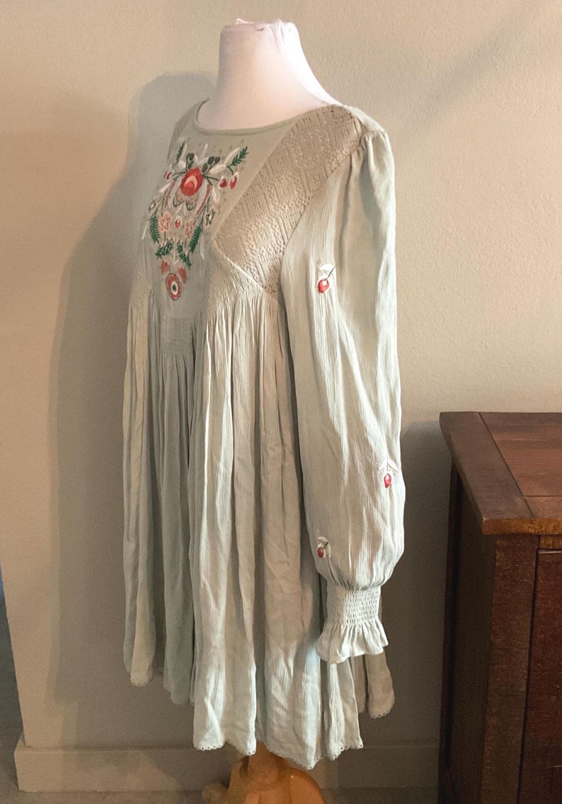 K&ouml;nnte beinhalten: Hellgr&uuml;nes Kleid mit floraler Stickerei und langen &Auml;rmeln. Das Kleid hat einen runden Ausschnitt und einen gerafften Rock. Die &Auml;rmel haben eine ger&uuml;schte Manschette und kleine rote Stickereiakzente. Das Kleid wird auf einer Schaufensterpuppe pr&auml;sentiert.