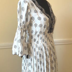 May include: White dress with a V-neck and floral pattern. The dress features bell sleeves, a cinched waist, and black and gold accents. The dress is displayed on a black mannequin.