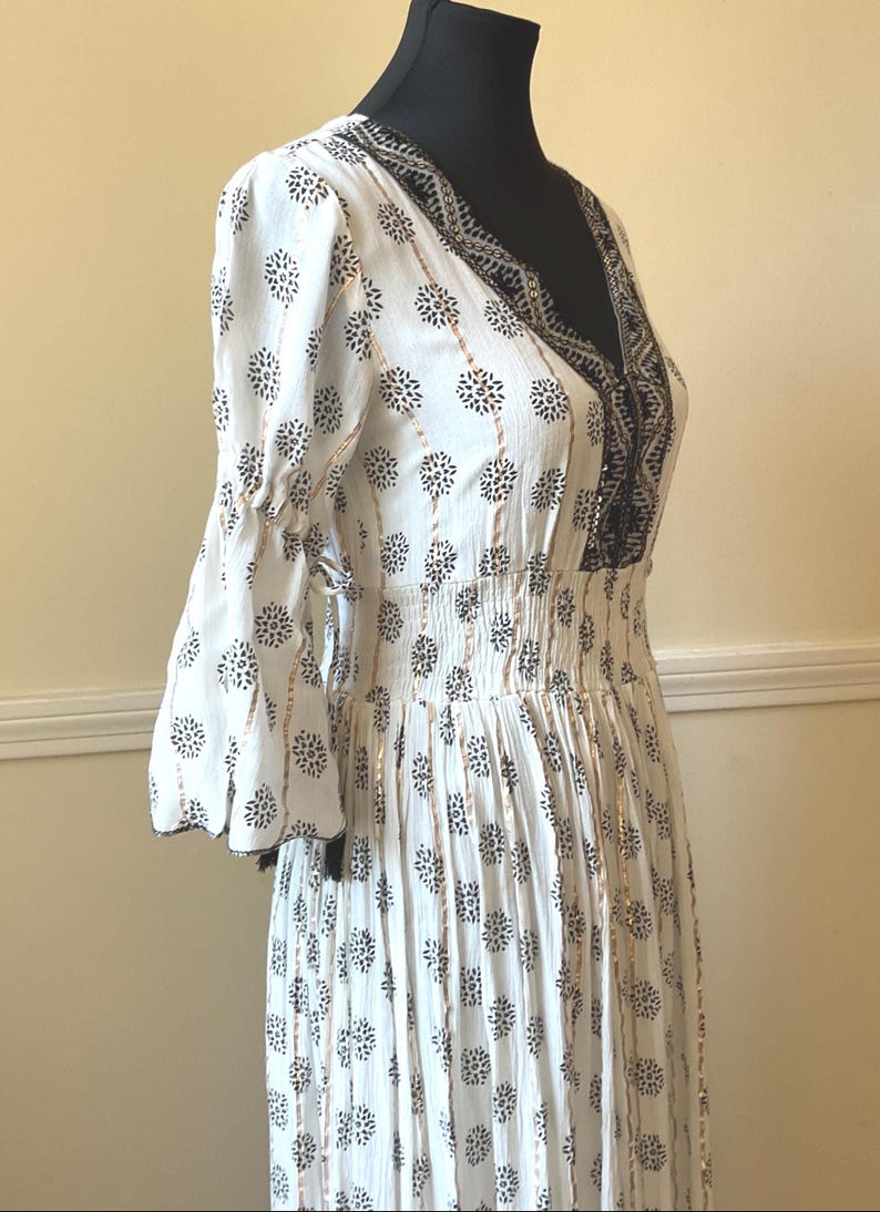May include: White dress with a V-neck and floral pattern. The dress features bell sleeves, a cinched waist, and black and gold accents. The dress is displayed on a black mannequin.