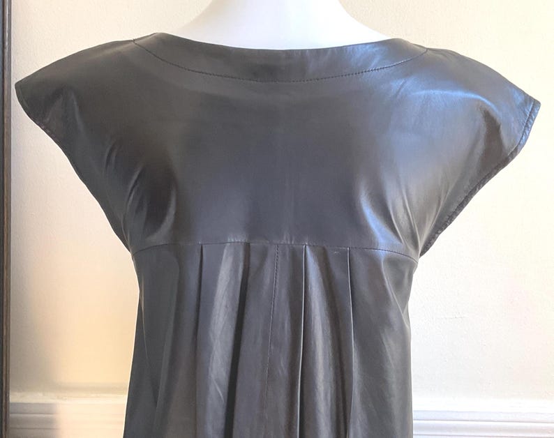 May include: A dark grey leather dress with a boat neck and cap sleeves. The dress has a pleated design below the bust and a subtle sheen. The dress is displayed on a mannequin.